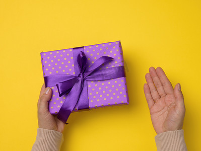 两只女手拿着黄色背景上的紫色礼盒，生日快乐的概念