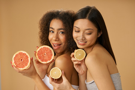 两位可爱的混血年轻女性的肖像，她们拿着葡萄柚和柠檬切成两半，同时在米色背景下摆出孤立的姿势