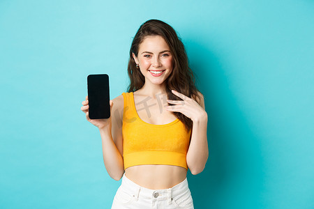 身穿夏装的时尚白人女性展示着空荡荡的智能手机屏幕，微笑着展示应用程序或在线商店，站在蓝色背景上