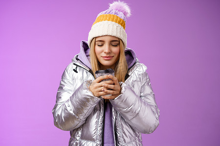 迷人温柔浪漫的金发女孩嗅着好闻的气味温暖美味的咖啡拿着外卖杯温暖的手闭上眼睛看起来很高兴戴着冬季夹克帽享受假期，紫色背景