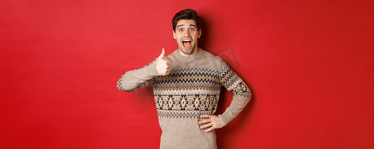 穿着圣诞毛衣的兴奋、有魅力的男人的肖像，竖起大拇指，看起来很惊讶，推荐好东西，为圣诞相关产品做广告