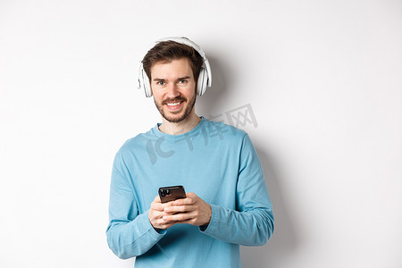 黑色摄影照片_迷人的微笑男人戴着无线耳机听音乐，使用黑色智能手机，看起来很高兴，白色背景