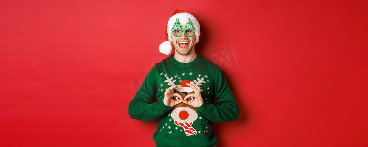 戴着圣诞帽和派对眼镜的无忧无虑的帅哥的画像，取笑他的圣诞毛衣，在红色背景下看起来很开心