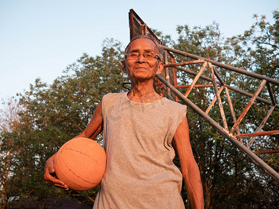 室外篮球场摄影照片_夏日，穿着运动服的亚洲老年男子拿着篮球，在室外篮球场看着镜头。