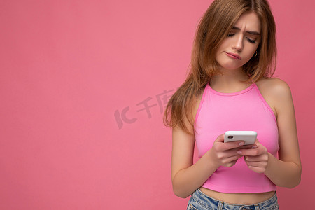 美丽体贴的年轻金发女性的特写肖像，她身穿粉红色上衣，在粉红色背景中与世隔绝，手握空白空间，使用手机上网上网，看着 gadjet 显示器并产生怀疑