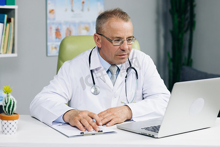 穿着白色医用外套和眼镜的中年高级主治医生坐在工作场所，与病人交谈，在笔记本电脑上进行视频通话，在纸质杂志上写笔记