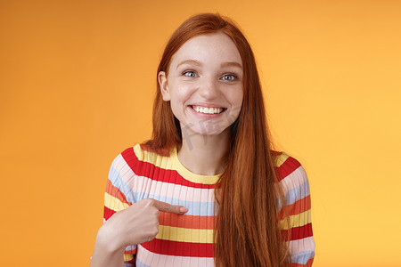 感恩钜惠倾情回馈摄影照片_快乐、兴奋、咧嘴笑的红发女孩选择了微笑的感激之情，高兴地指着自己，看起来很惊讶，感恩的相机找到了工作，获得了站在橙色背景下的奖学金