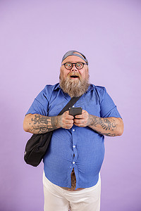 带斜挎包和眼镜的风趣大个子拿着紫色背景的手机