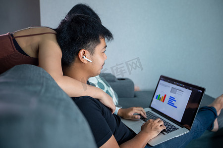 亚洲年轻微笑夫妇拥抱并使用无线耳机，并使用在家工作的电脑笔记本。他正在为在家的在线会议准备图表和信息。
