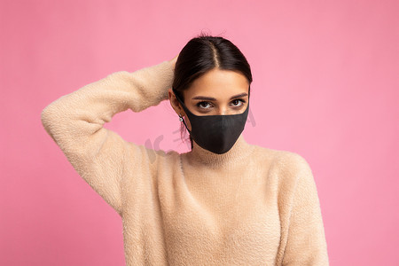 年轻美丽的褐发女性，脸上戴着可重复使用的病毒防护面具，抵御粉红色背景墙上分离出的冠状病毒