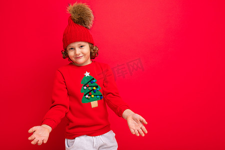 穿着红色圣诞毛衣的酷少年在红墙、温暖的帽子和带圣诞树的毛衣的背景下闲逛