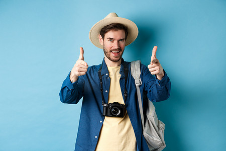 带你去旅游摄影照片_帅气的游客带着相机和背包去暑假，用手指指着相机，站在蓝色背景上邀请你