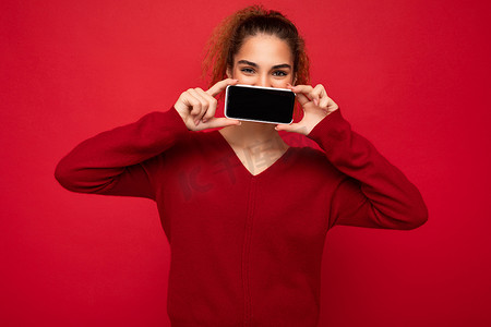 特写照片：快乐风趣的年轻女性穿着深红色毛衣，在红色背景中被隔离，拿着智能手机，并显示手机屏幕，上面有复制空间，可以看到相机