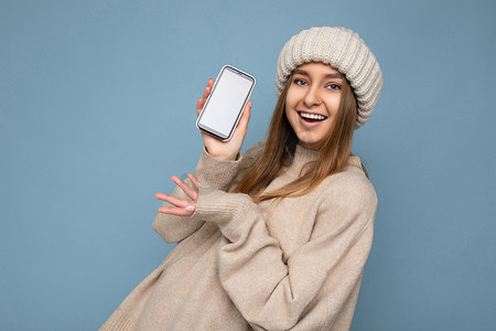 照片中，美丽微笑、正面漂亮的年轻女性身穿时尚的米色毛衣，头戴米色针织冬帽，在蓝色背景中与世隔绝，手握空白空间，展示手机，屏幕空空如也，供模拟观看