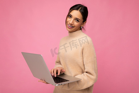 职业照摄影照片_照片中，穿着米色毛衣的美丽微笑快乐的黑发年轻女子拿着上网本电脑在键盘上打字，看着粉红色墙壁背景上孤立的相机