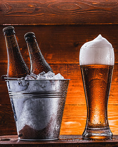 瓶啤摄影照片_两瓶啤酒装在一个桶里，里面装着冰，还有一杯啤酒，泡沫丰富，紧挨着深色背景
