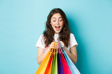 快乐的年轻女人兴奋地看着她的购物袋，在商店打折购买，站在蓝色背景中