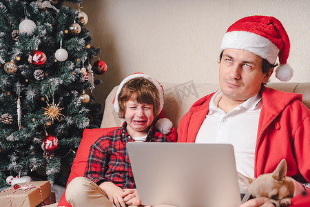 戴着圣诞帽的家庭、父亲和哭泣的男孩，带着笔记本电脑和狗坐在客厅的沙发上