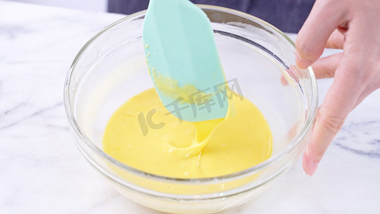 蛋黄摄影照片_用绿色橡胶抹刀混合器工具将蛋黄混合到蛋糕糊中，搅拌直至光滑并在玻璃碗中充分混合，特写，生活方式
