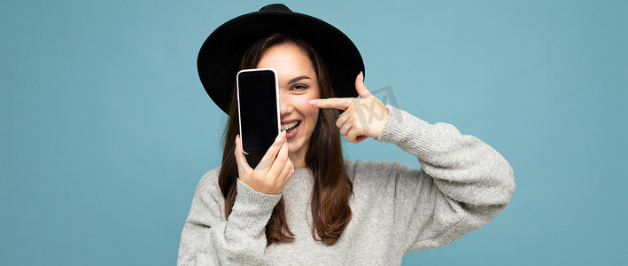 戴着黑色帽子和灰色毛衣的美丽积极女性手持手机的全景照片，显示智能手机在背景中被隔离，看着相机