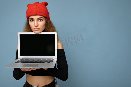 特写肖像的滑稽美丽的深色金发女人拿着笔记本电脑看着相机穿着黑色裁剪上衣和红色和橙色的 do-rag 隔离在浅蓝色的墙壁背景