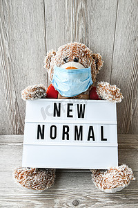 泰迪熊玩偶戴着面罩背包，手拿灯箱，木质背景上写着 NEW NORMAL 文字，复制空间，2020 年隔离。回到学校
