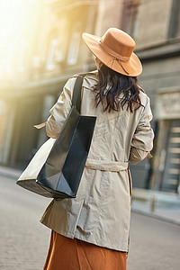 一位穿着秋灰色外套和帽子、大购物袋走在城市街道上的时尚女性的后视