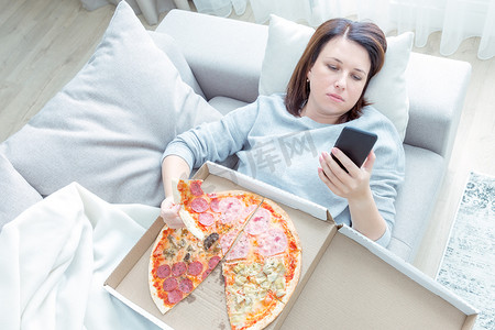 悲伤的女人吃披萨，拿着手机躺在家里的沙发上，蓝色调