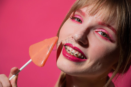 一位年轻女子的画像，她戴着牙套，妆容鲜艳，在粉红色的背景下吃着棒棒糖。