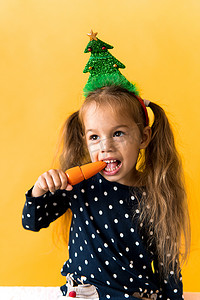 小鹿女孩摄影照片_肖像正面开朗微笑快乐的小女生女孩圣诞树装饰波尔卡圆点连衣裙咬吃橙色背景上的橙色胡萝卜。