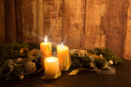 圣诞节概念的温暖：三根蜡烛点亮，十字屏幕星星在一张深色木桌上产生效果，桌子上有松枝、天然松果、金色缎带和白色欧根纱丝带