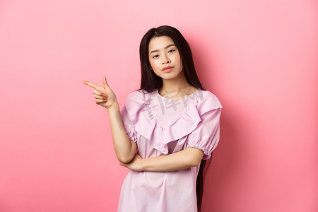 自鸣得意摄影照片_年轻自鸣得意的亚洲女孩看起来很酷，手指留在标志上，在粉红色浪漫背景下做广告产品