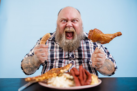 情绪化的肥胖男人拿着香肠和熏鸡腿坐在桌边