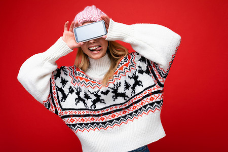 照片中，金发美女微笑着，头戴温暖的针织帽，身穿冬日暖和的毛衣，在红色背景中与世隔绝，展示的智能手机屏幕空空如也，可以玩得开心，也可以展示舌头