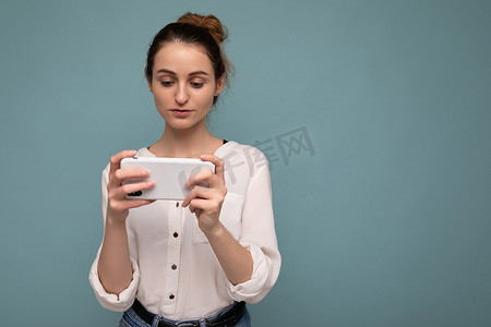 穿着休闲服的漂亮年轻女性站在孤立的背景中，通过手机看手机屏幕在互联网上冲浪