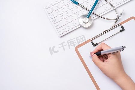 女医生在白色工作台上用听诊器、电脑键盘在剪贴板上写病历。
