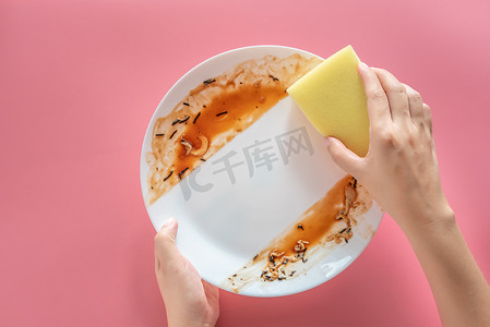 妇女在吃完粉色背景上隔离的饭后，用黄色清洁海绵清理和清洗白色盘子上的食物污渍和污垢。