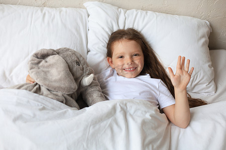 可爱的大象摄影照片_可爱的小女孩醒来，躺在床上，手里拿着一只玩具大象。