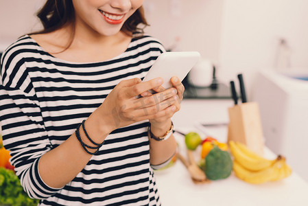 app纯摄影照片_迷人的年轻女孩在厨房里穿着围裙忙着使用手机或检查智能手机的更新