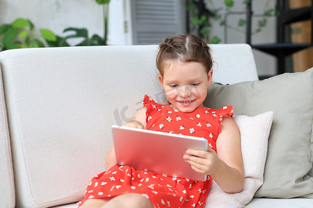 微笑的小女孩躺在沙发上玩在线游戏，网上冲浪信息，在平板电脑上使用有趣的应用程序。