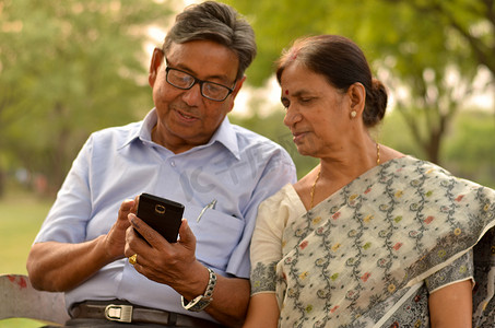 印度新德里摄影照片_印度新德里，一对老夫妇坐在公园长椅上，看着智能手机，笑着，重点放在手上
