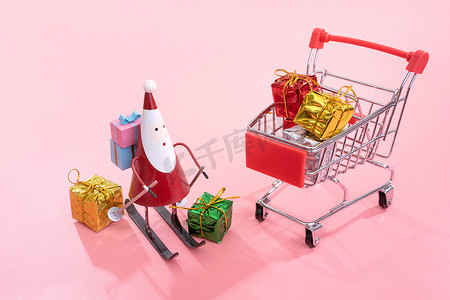 礼盒元素摄影照片_圣诞购物概念、带圣诞老人玩具的迷你红色购物车手推车和淡粉色背景中突显的礼盒、空白复制空间、特写