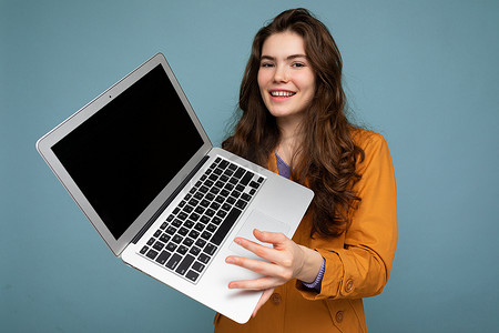 照片中美丽微笑的快乐年轻女子拿着电脑笔记本电脑看着相机，穿着与墙壁背景隔离的休闲智能服装，玩得很开心