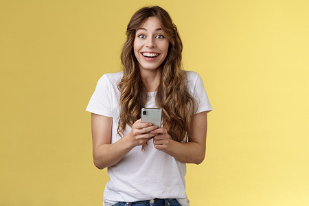非常高兴热情惊讶快乐可爱的女性女孩在网上收到令人愉快的感人消息拿着智能手机广泛微笑看着相机快乐有趣的黄色背景