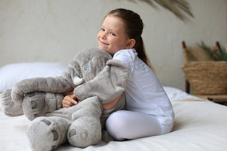 可爱的大象摄影照片_可爱的小女孩坐在家里的床上，带着玩具大象。