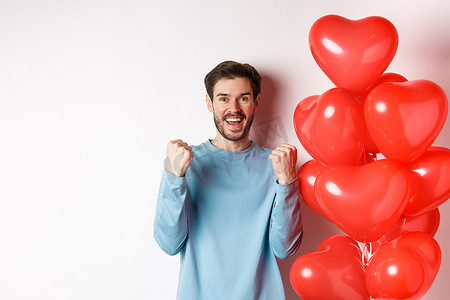肯定摄影照片_庆祝情侣日的快乐男人的肖像，站在情人节红心气球旁欢呼，做出肯定的手势，对着镜头微笑，站在白色背景上