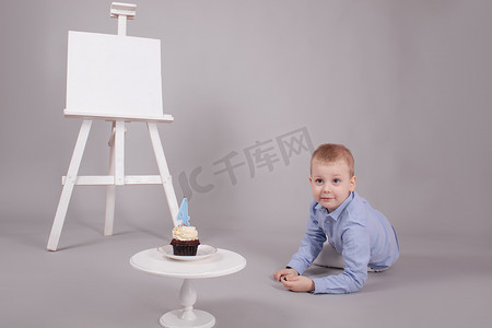带蜡烛蛋糕摄影照片_学龄前男孩穿着白色裤子和蓝色衬衫，灰色背景，靠近画架，上面有模型和带蜡烛的纸杯蛋糕，形状为四号 4。生日快乐。