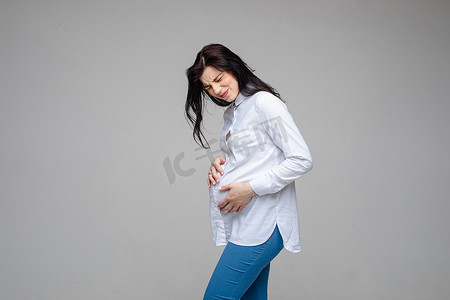 母性摄影照片_美丽的女性怀孕了，在灰色的工作室背景下触摸怀孕的腹部感觉疼痛。