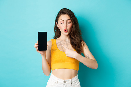 夏季，年轻兴奋的女性穿着短上衣，指着并看着空荡荡的智能手机屏幕，展示应用程序或网上商店，站在蓝色背景上