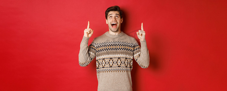 喜欢寒假的兴奋帅哥展示广告，惊讶地看着并用手指指着，站在红色背景的圣诞毛衣上